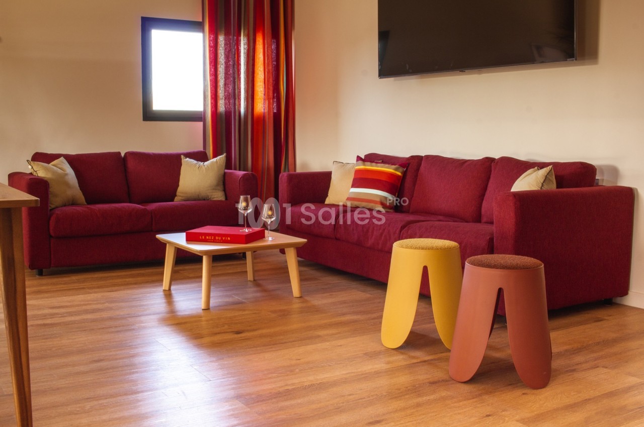 Salon avec deux canapés rouges, table basse en bois, tabourets colorés, rideaux rouges et télévision murale.