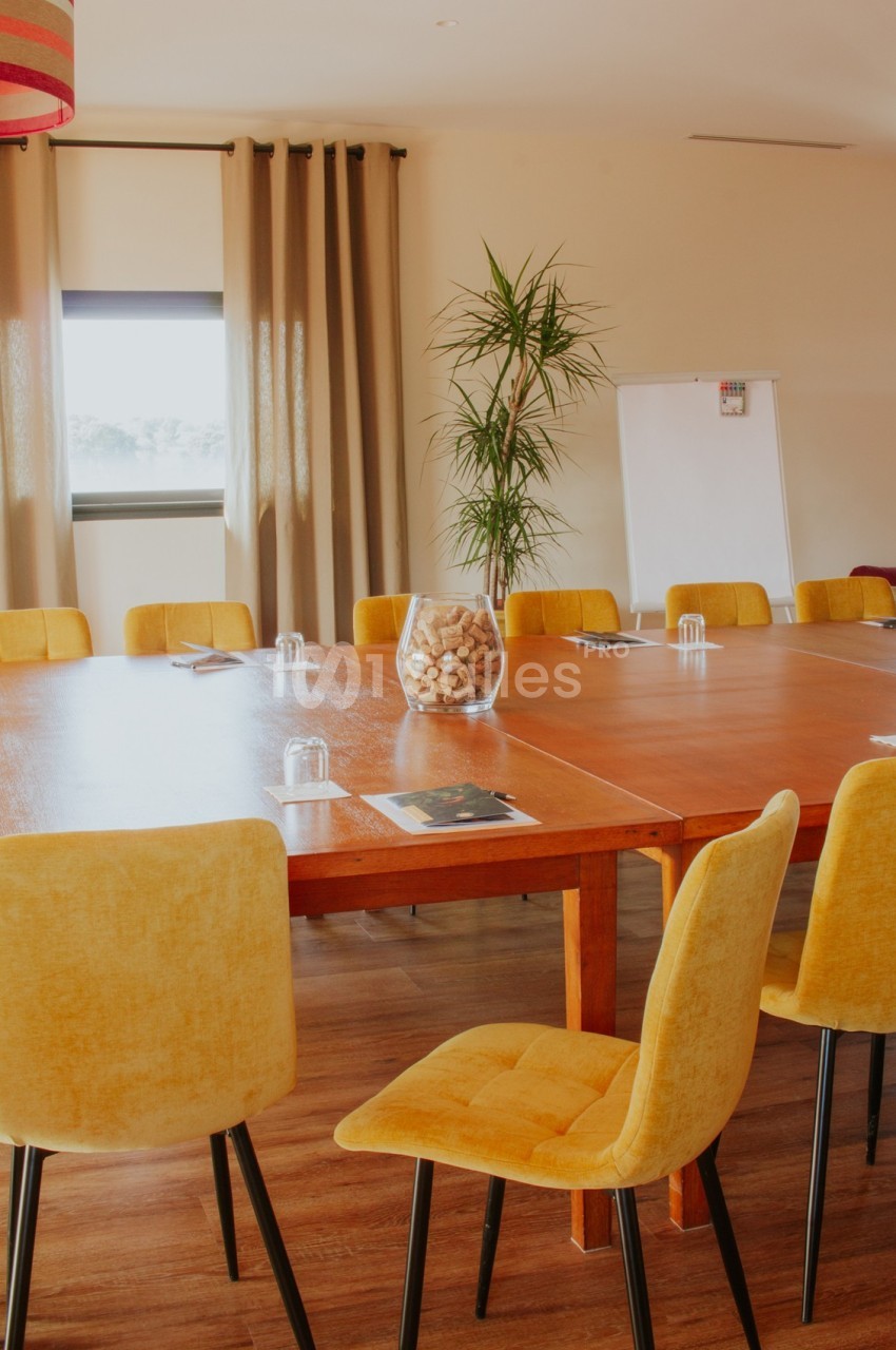 Salle de réunion lumineuse avec grande table en bois, chaises jaunes, tableau blanc et plante en arrière-plan.
