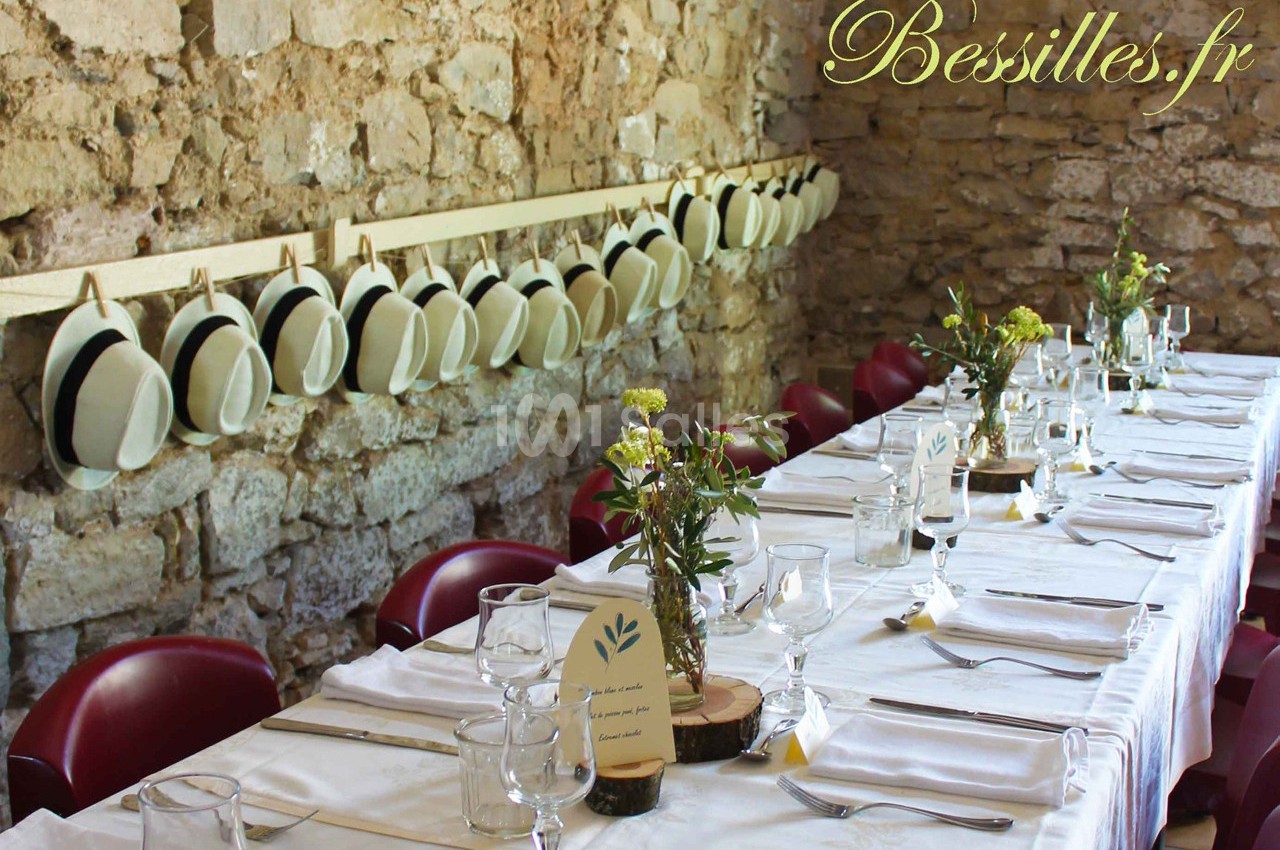 Table dressée avec nappes blanches, décor champêtre, chapeaux suspendus sur un mur en pierre.