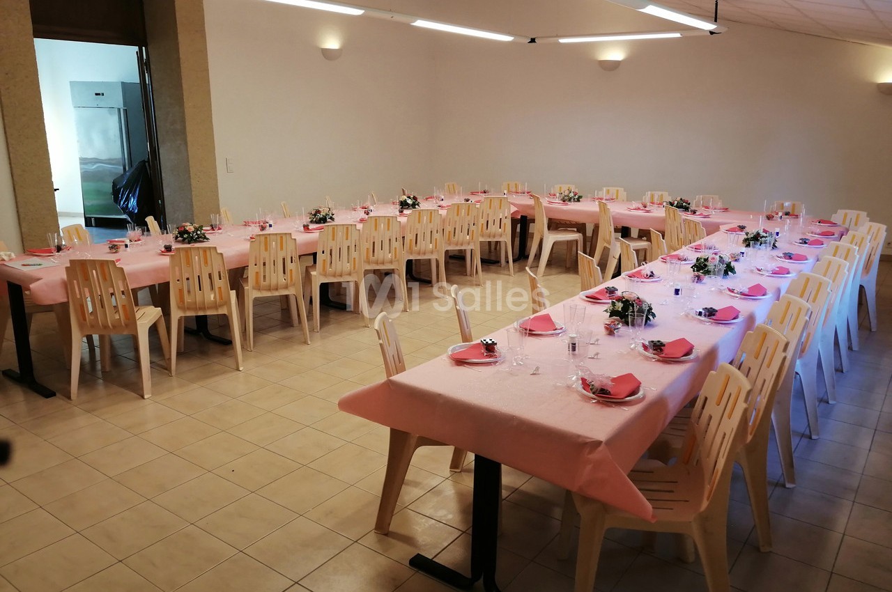 Salle décorée pour un repas, avec des tables rectangulaires couvertes de nappes roses et des chaises en plastique beige.