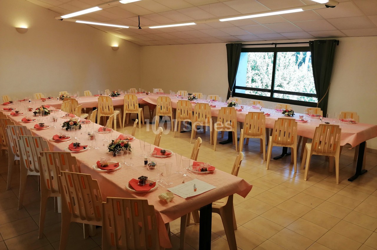 Salle décorée pour un repas, avec tables alignées, nappes roses, chaises en plastique et fenêtres donnant sur un extérieur…