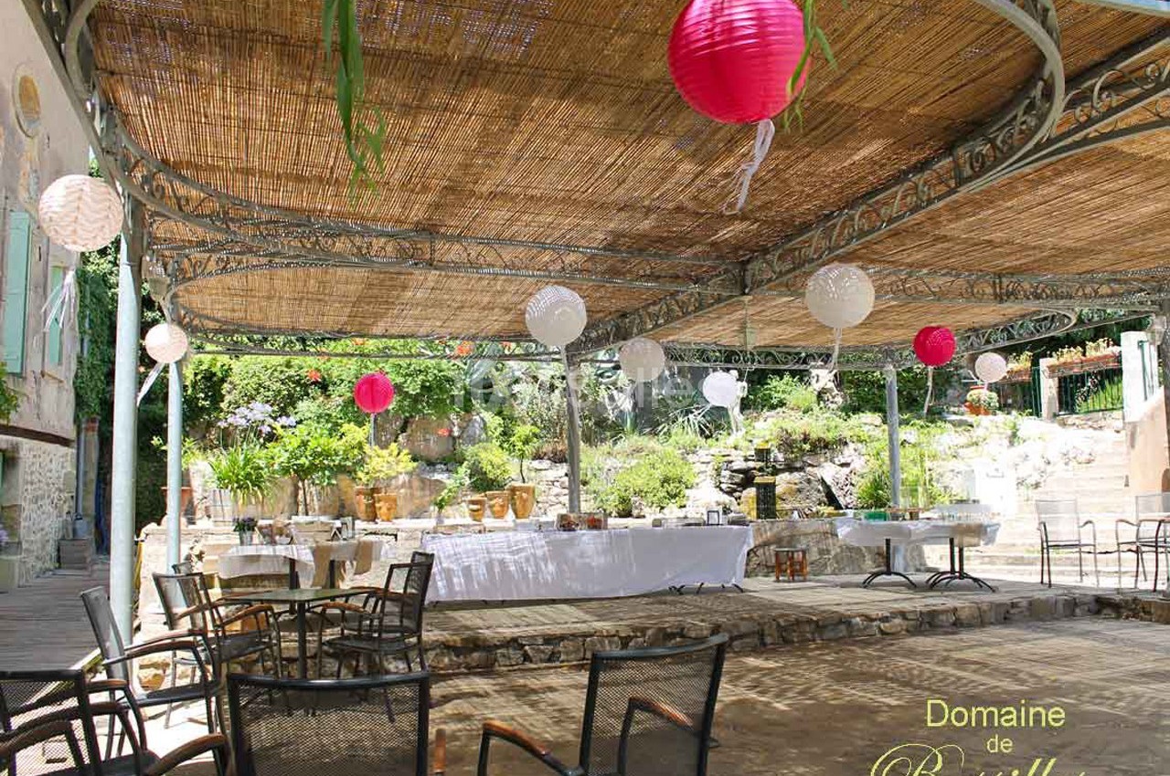 Terrasse ombragée avec tables dressées, lanternes suspendues et végétation en arrière-plan.
