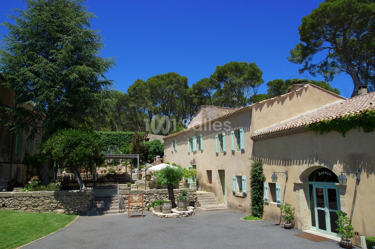 Cour d'une maison en pierre avec volets verts, entourée d'arbres et d'un ciel bleu dégagé.