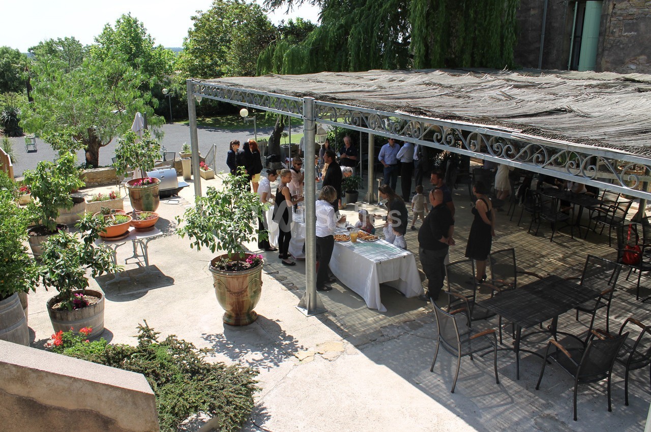 Des personnes rassemblées sous une pergola dans une cour extérieure, avec des tables et des plantes en pot.