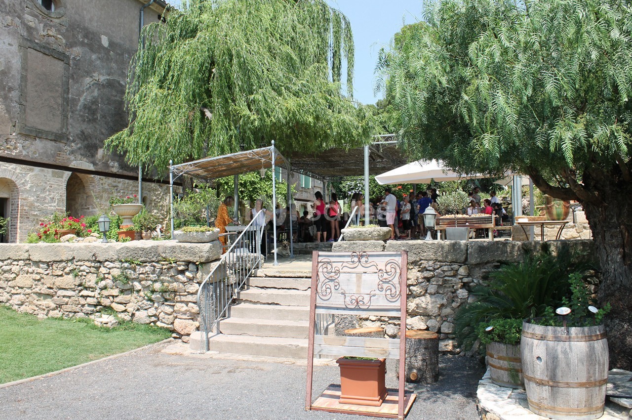 Terrasse ombragée d'un restaurant en plein air avec des tables occupées, entourée de végétation et de murs en pierre.