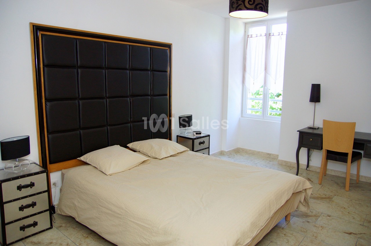 Chambre lumineuse avec lit double, tête de lit rembourrée, bureau en bois, lampes et fenêtre donnant sur l'extérieur.