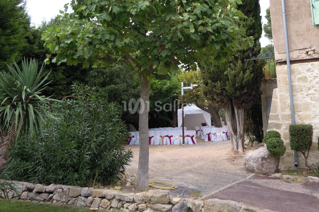 Chaises blanches décorées sous une tente dans un jardin arboré, près d'une maison en pierre.