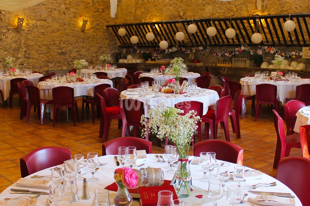 Salle de réception décorée avec des tables rondes dressées, chaises rouges et guirlandes lumineuses suspendues.