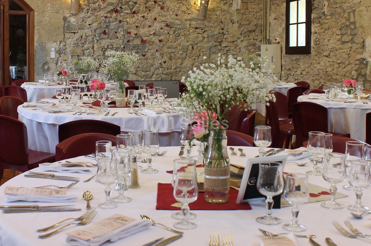Salle de réception avec des tables rondes dressées, décorées de fleurs et entourées de chaises rouges.