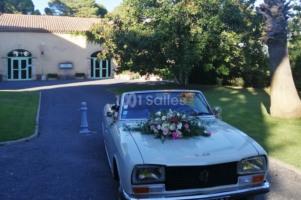 Voiture ancienne blanche décorée de fleurs, stationnée devant un bâtiment avec des arches et un jardin arboré.