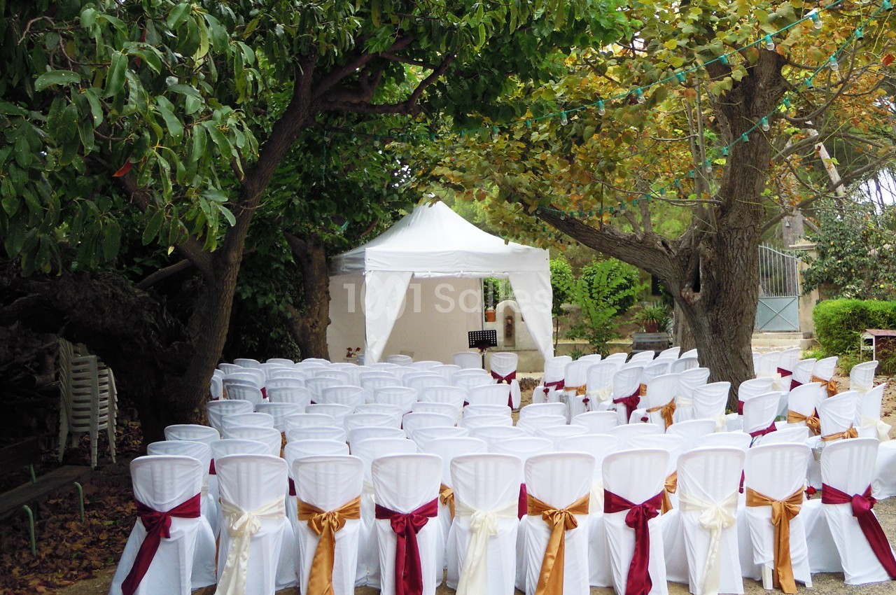 Chaises blanches décorées de rubans rouges et dorés disposées en rangées devant une tente blanche dans un jardin.