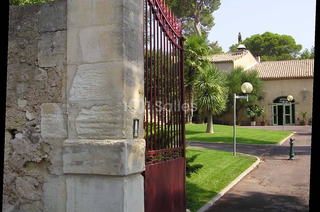 Portail rouge ouvert donnant sur une allée bordée de pelouse et menant à une maison entourée de végétation.