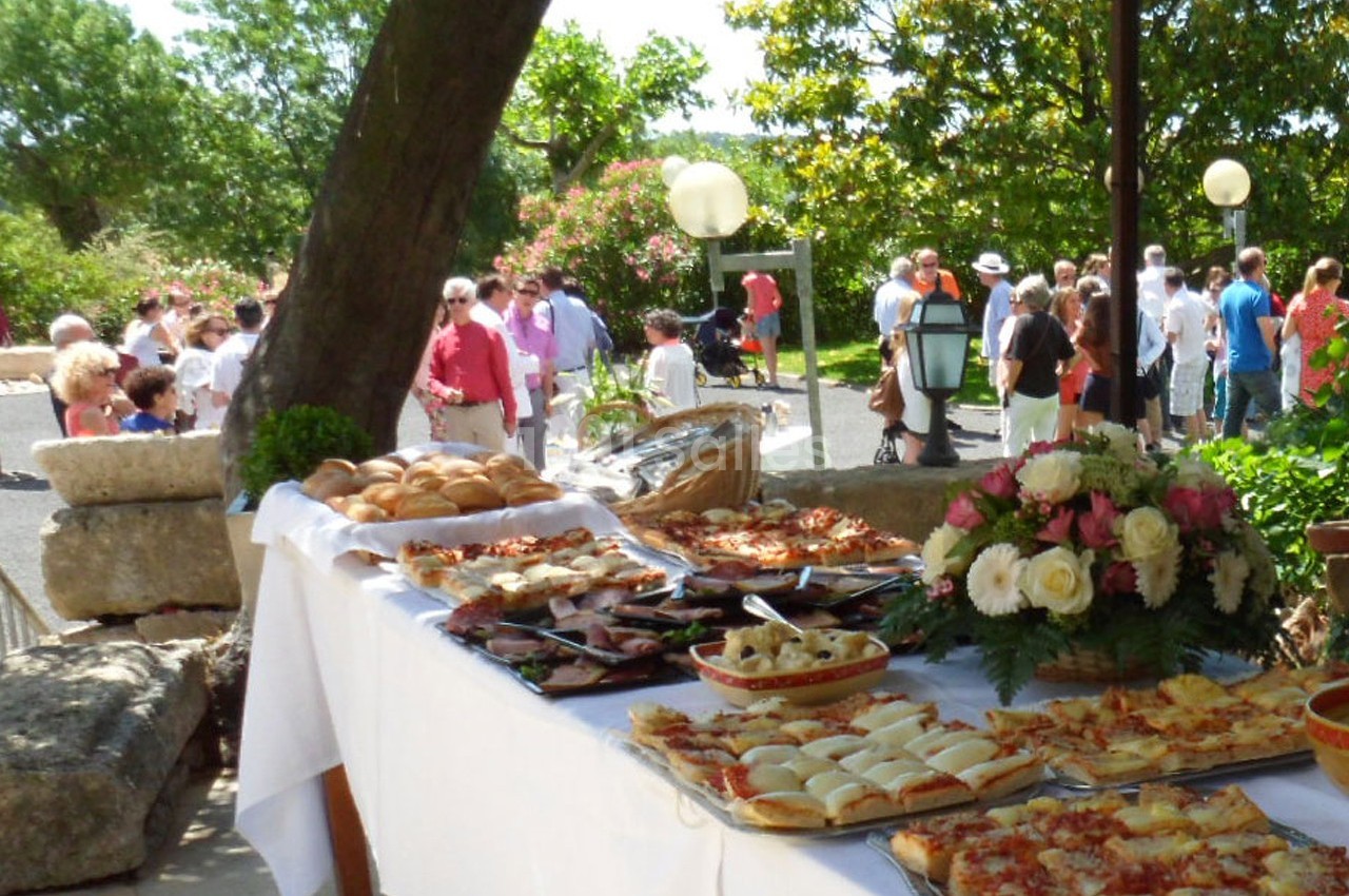 Buffet en plein air avec pizzas, pains et fleurs, entouré de convives dans un jardin ensoleillé.