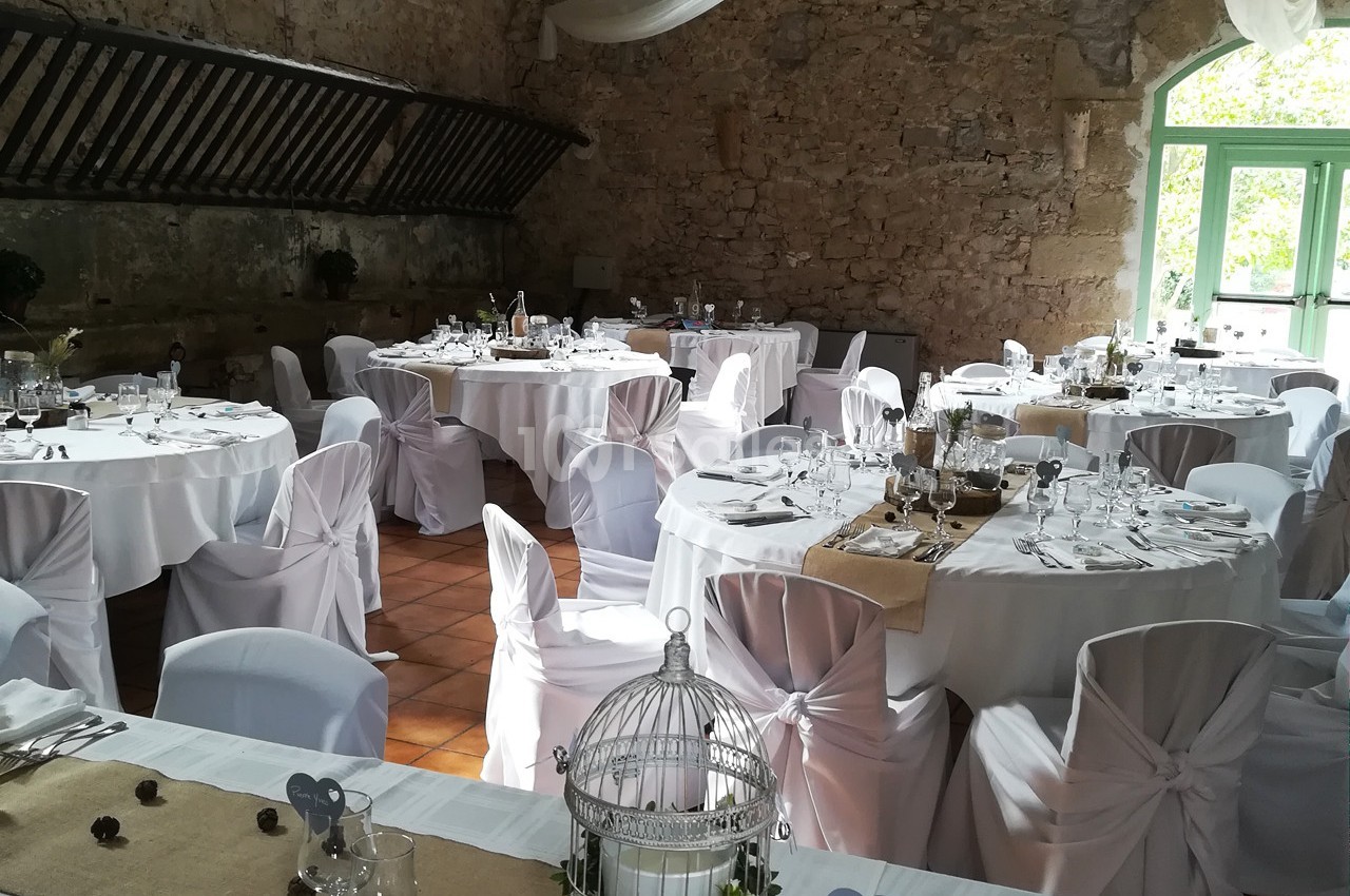 Salle de réception décorée avec des tables rondes dressées, nappes blanches et éléments rustiques dans un cadre en pierre.