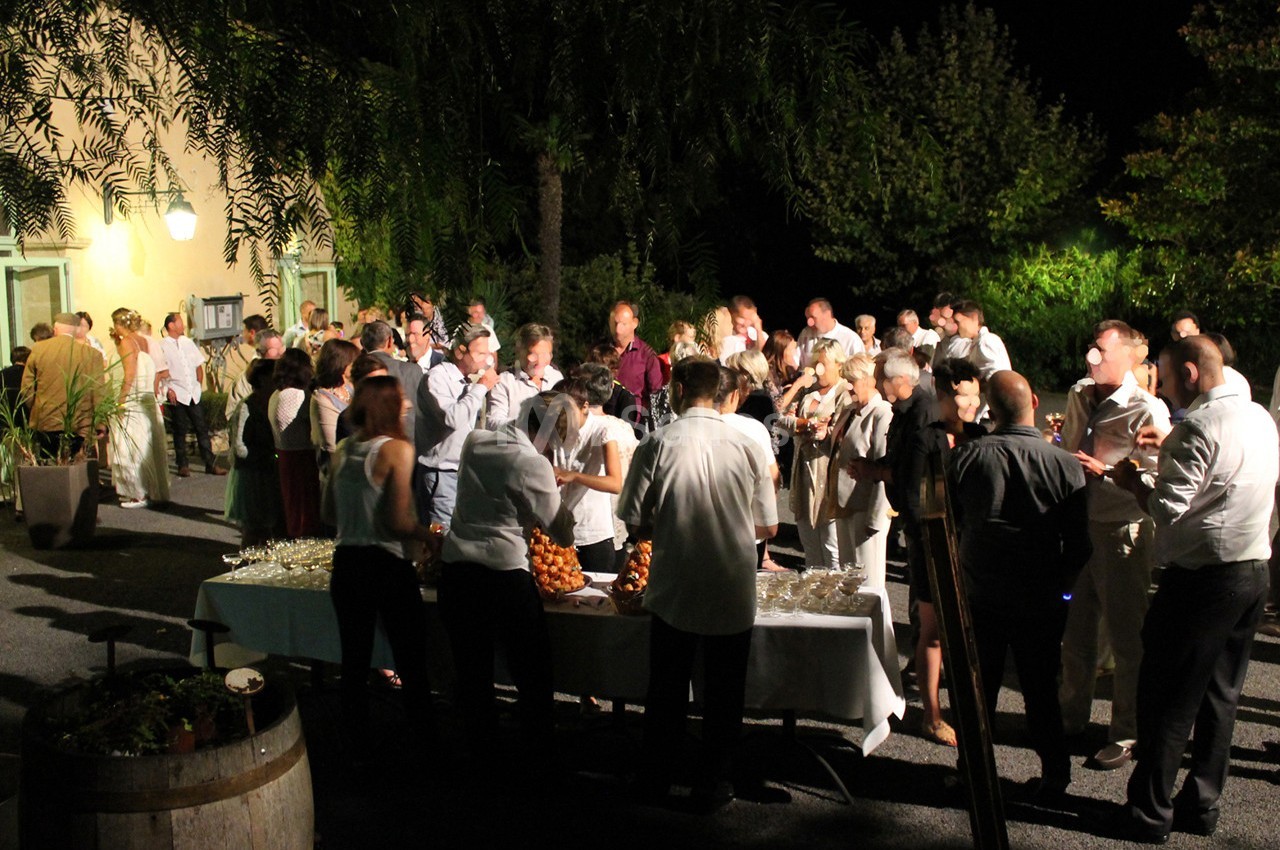Groupe de personnes rassemblées en soirée autour d'une table de buffet en extérieur, éclairée par des lumières.
