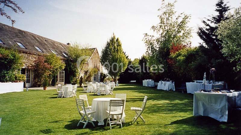 Jardin avec pelouse, tables et chaises blanches disposées pour un événement en plein air, entouré de verdure et d'arbres.
