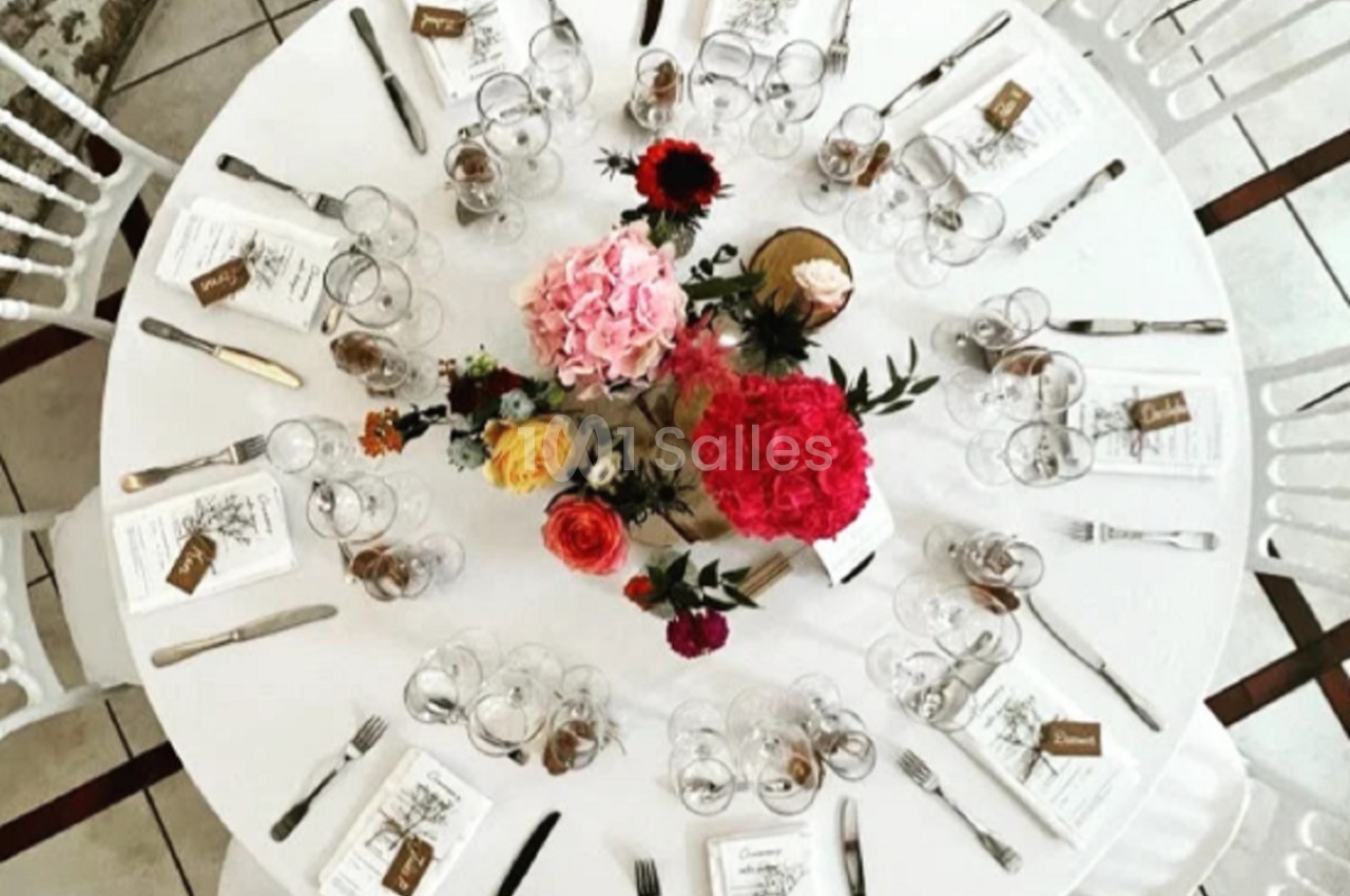 Table ronde dressée avec vaisselle, couverts, menus et centre de table floral coloré, entourée de chaises blanches.