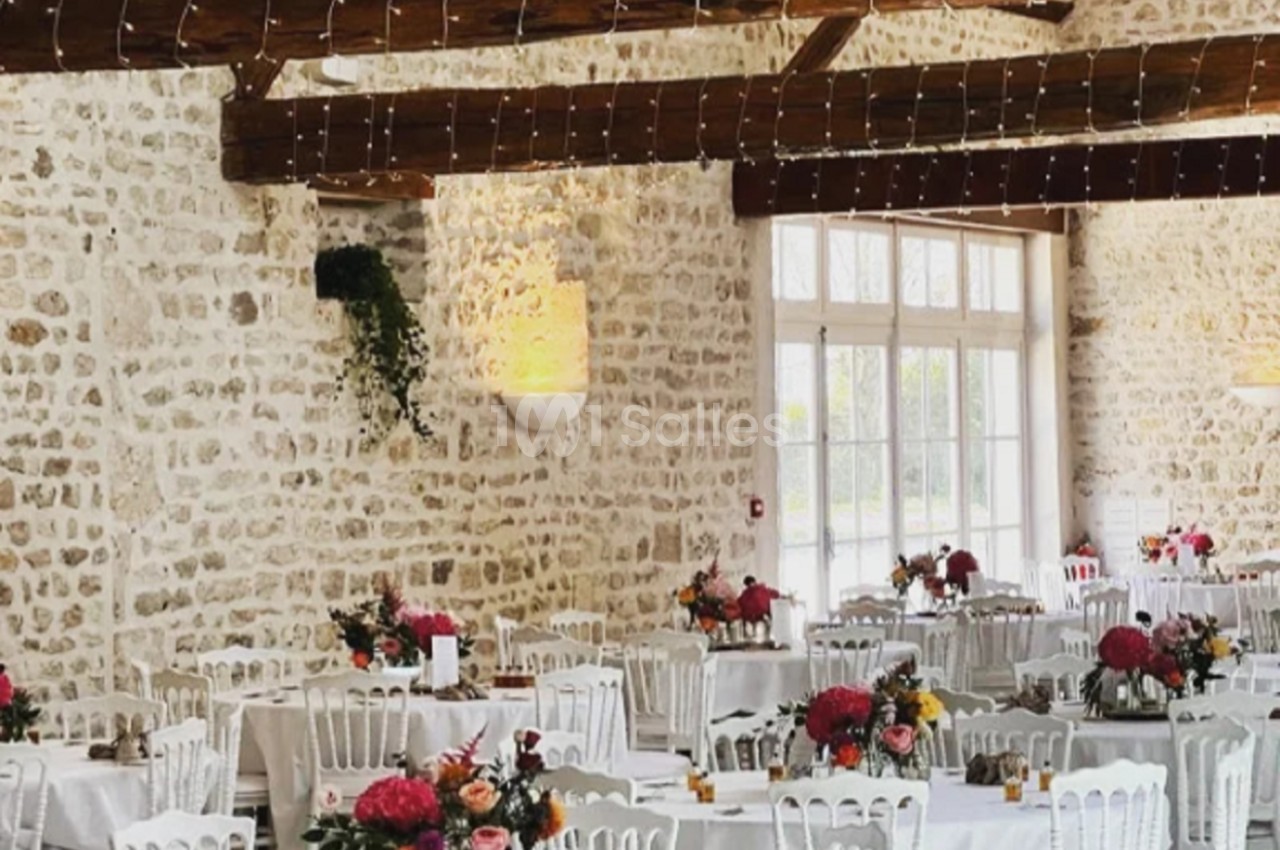 Salle de réception avec murs en pierre, tables rondes décorées de fleurs et chaises blanches sous des poutres apparentes.