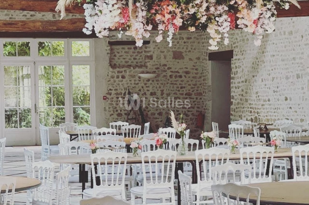 Salle de réception lumineuse avec tables rondes, chaises blanches et une décoration florale suspendue au plafond.