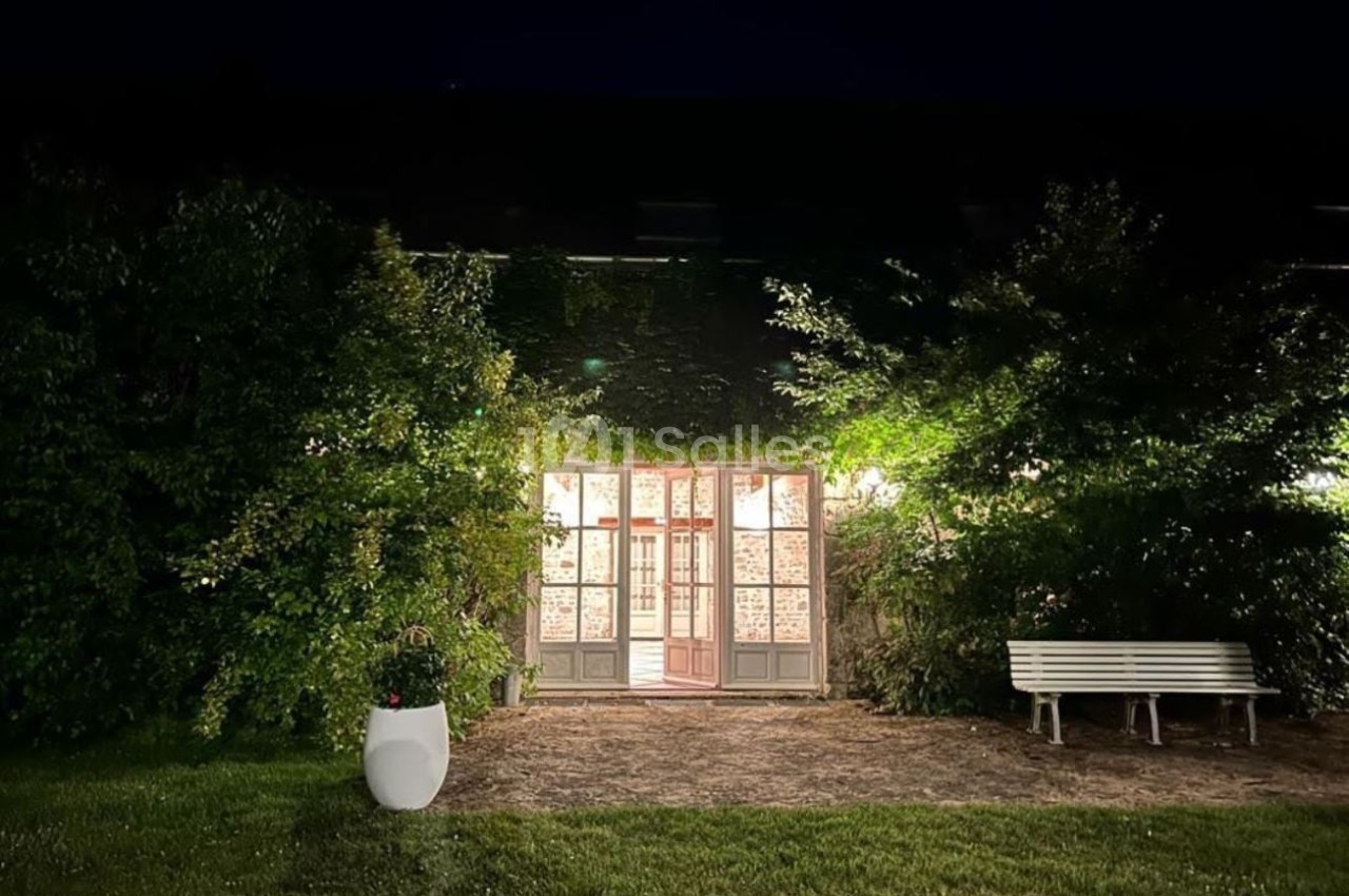 Façade vitrée éclairée entourée de végétation, avec un banc blanc et un pot de fleurs au premier plan, de nuit.
