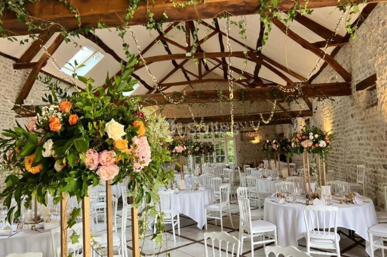 Salle de réception lumineuse avec poutres apparentes, tables décorées de nappes blanches et compositions florales colorées.