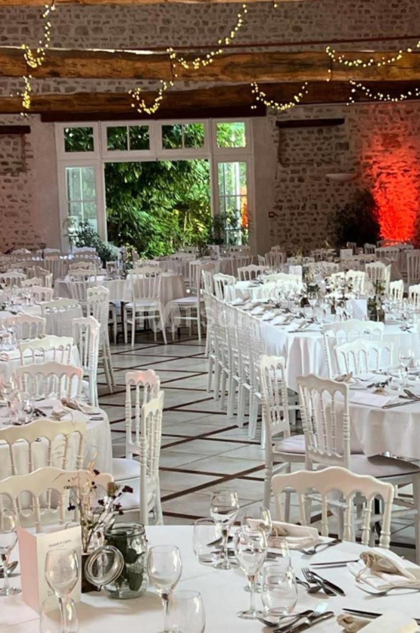 Salle de réception décorée avec des tables rondes dressées, chaises blanches et guirlandes lumineuses suspendues.