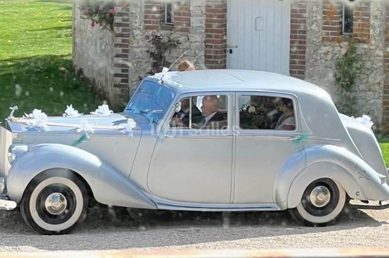 Voiture ancienne argentée décorée pour un mariage, stationnée devant un bâtiment en pierre avec pelouse en arrière-plan.