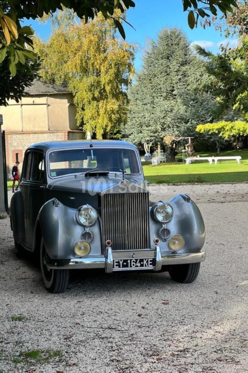 Voiture ancienne grise stationnée sur un chemin gravillonné, entourée d'arbres et d'un bâtiment en arrière-plan.