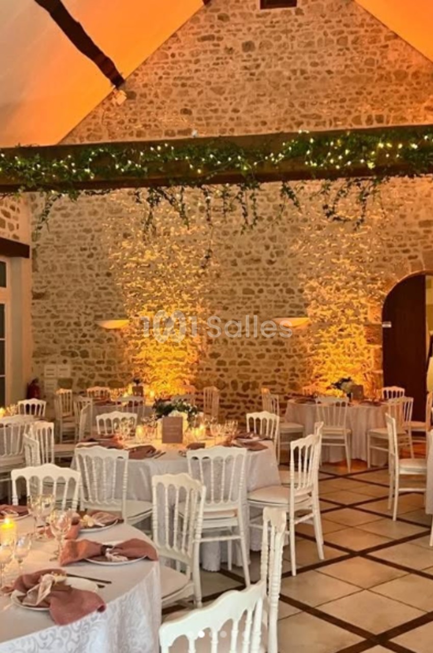 Salle de réception en pierre avec tables décorées, chaises blanches et guirlandes lumineuses suspendues.