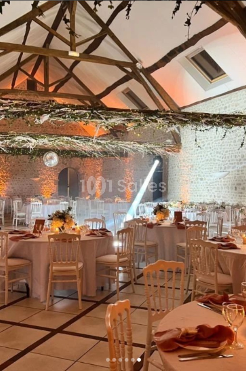 Salle de réception décorée avec des tables rondes, nappes blanches, guirlandes lumineuses et poutres apparentes.