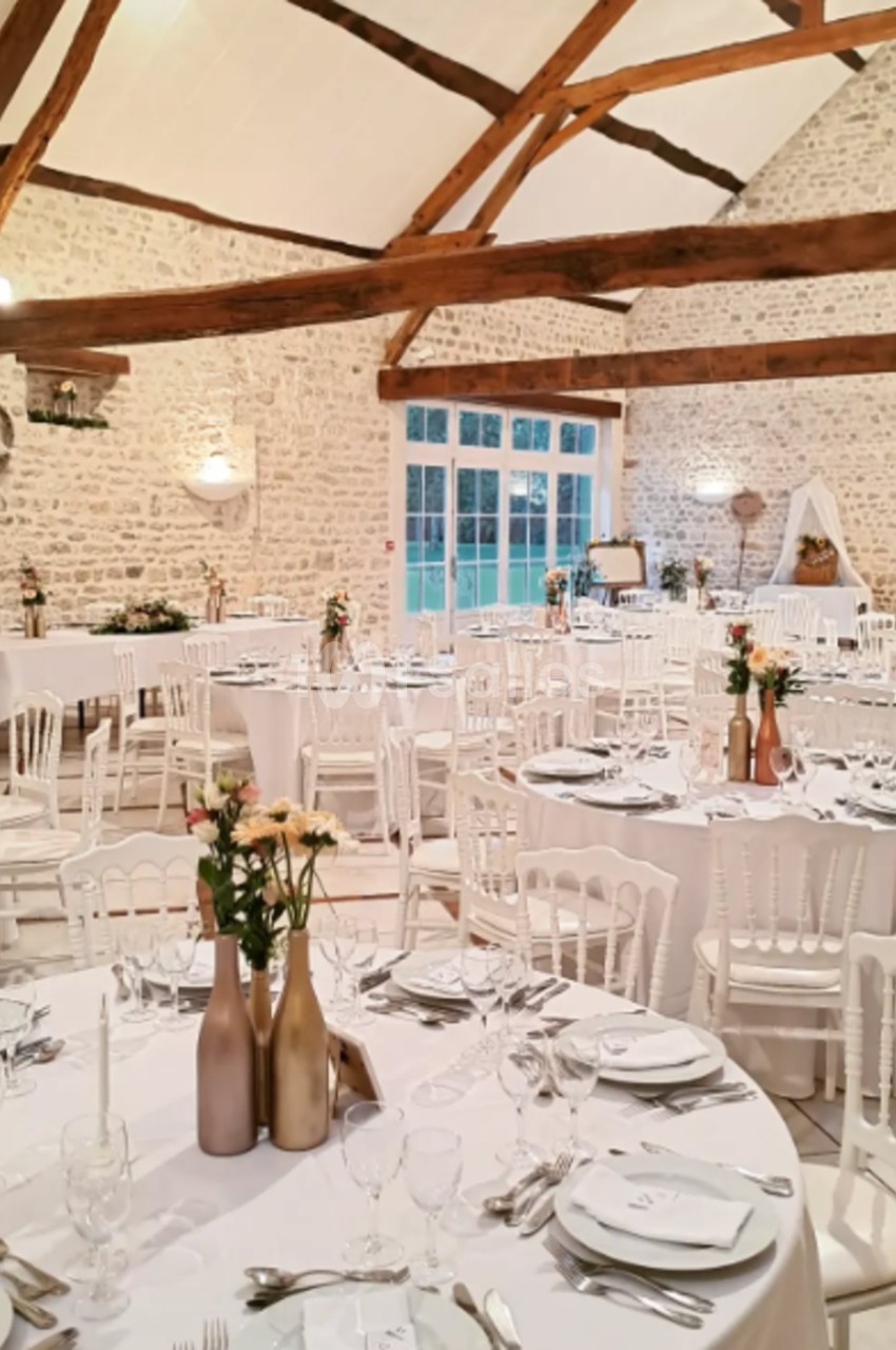 Salle de réception lumineuse avec murs en pierre, poutres apparentes et tables élégamment dressées pour un événement.