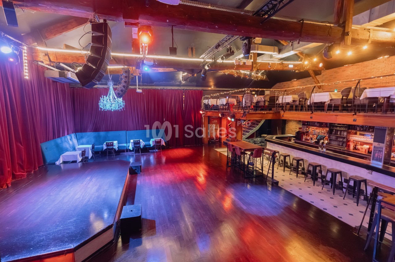 Salle spacieuse avec scène, parquet, rideaux rouges, bar, tables et mezzanine dans une ambiance lumineuse chaleureuse.