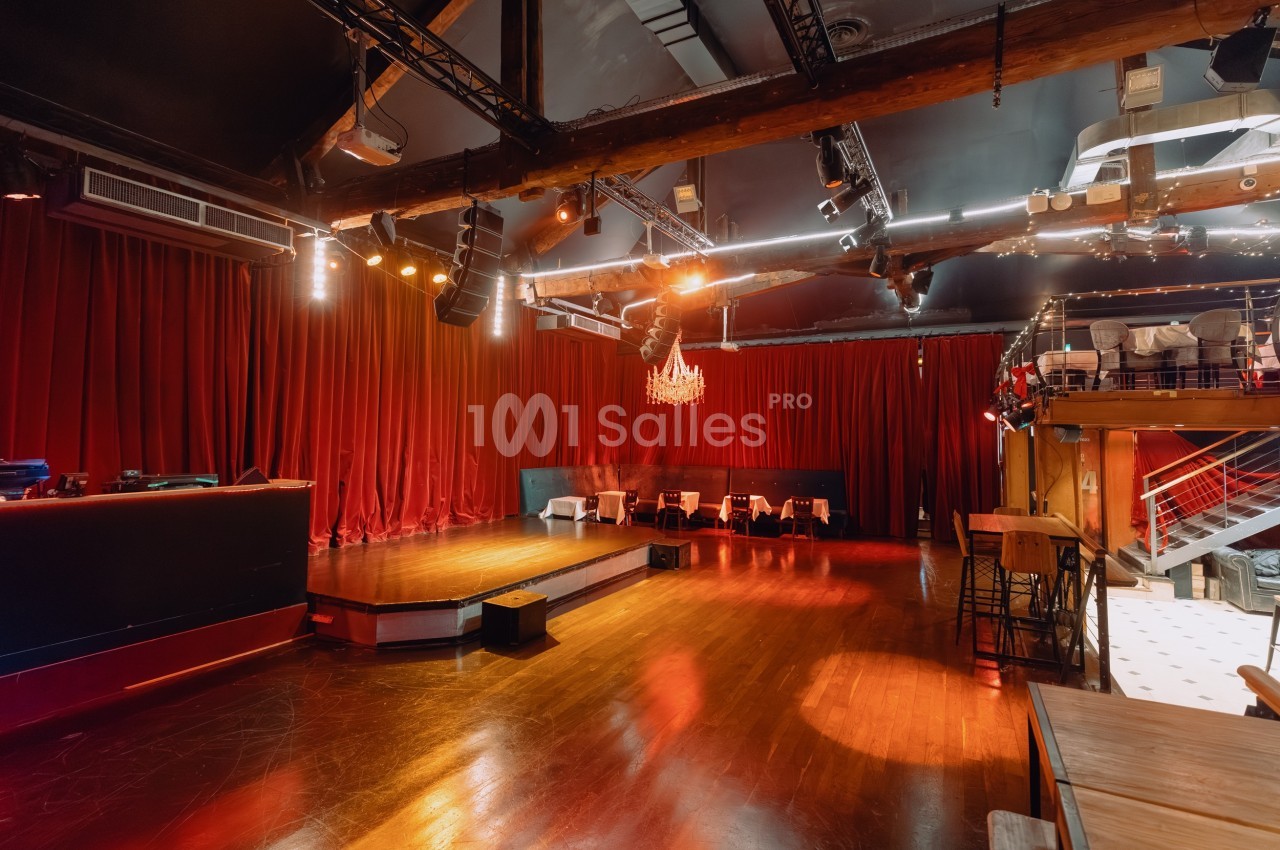 Salle de spectacle vide avec scène en bois, rideaux rouges, éclairage tamisé et mobilier disposé autour.