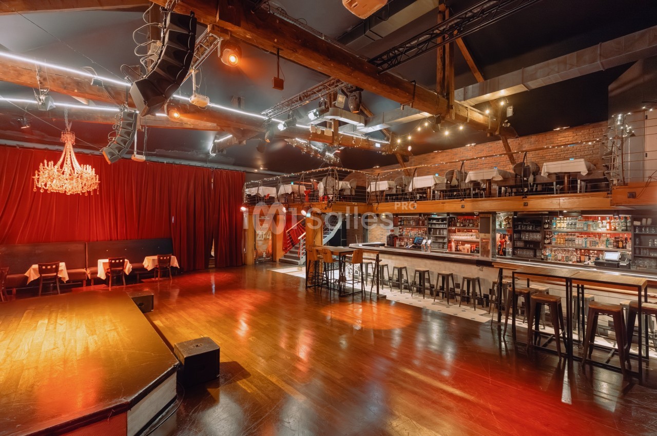 Salle de spectacle avec scène, bar, tables en mezzanine, éclairage chaleureux et décorations en bois et briques.
