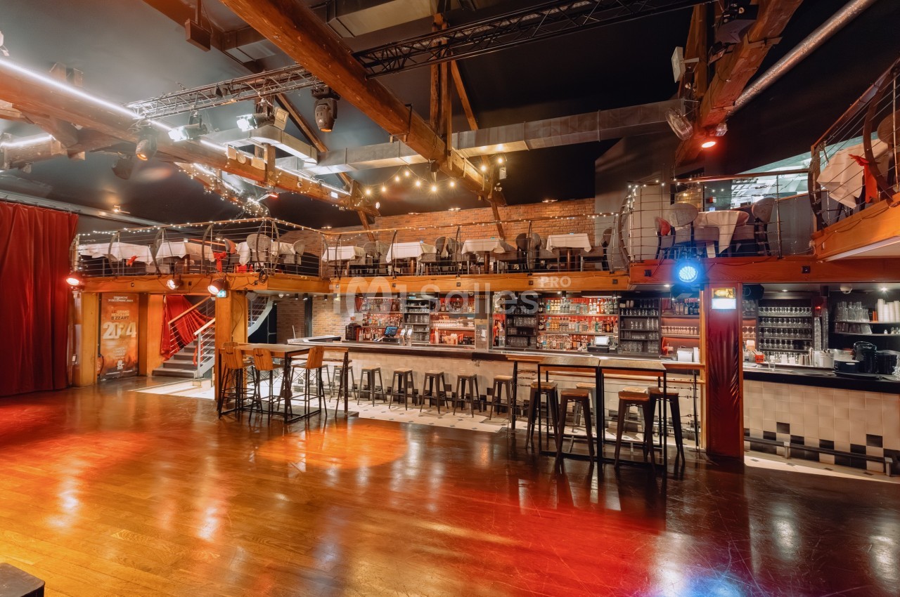Salle avec un bar central, tabourets alignés, mezzanine avec tables, éclairage chaleureux et poutres apparentes.