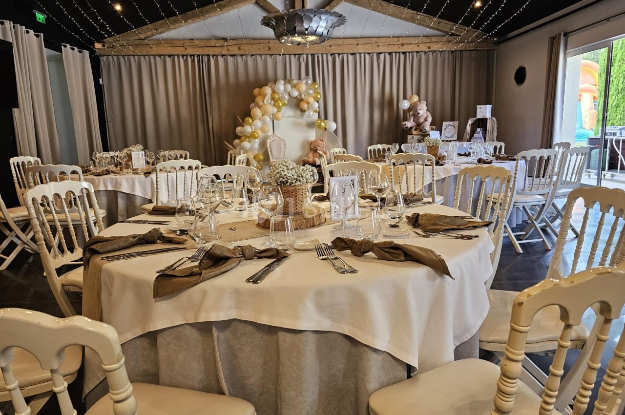 Salle décorée pour un événement avec des tables rondes dressées, des chaises blanches et des ballons en arrière-plan.