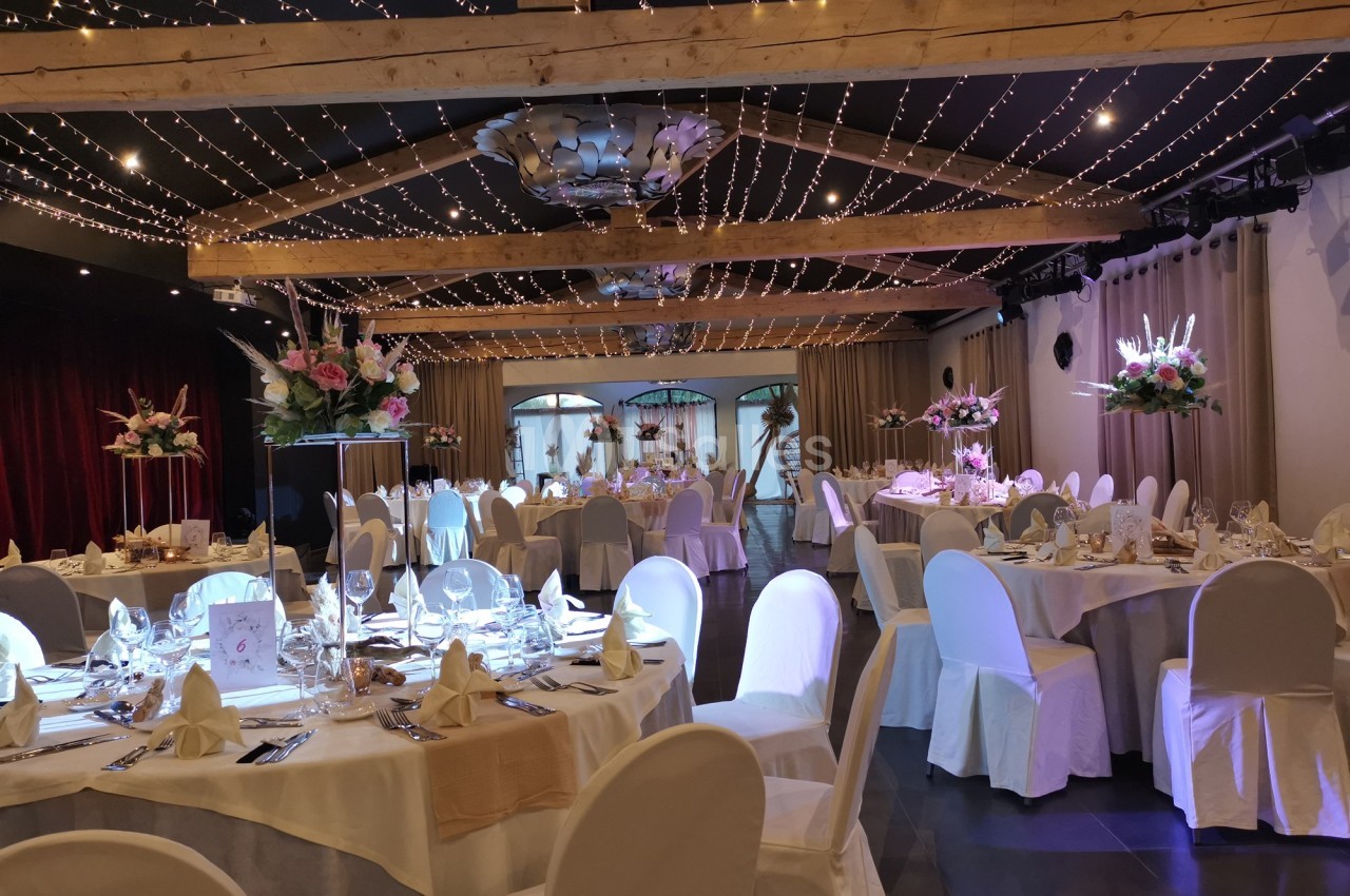 Salle de réception élégamment décorée avec des tables rondes, nappes blanches, chaises houssées et guirlandes lumineuses au…