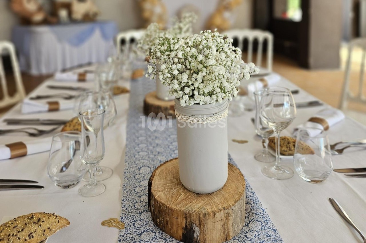 Centre de table avec un vase blanc rempli de gypsophiles, posé sur une rondelle de bois, entouré de couverts dressés.