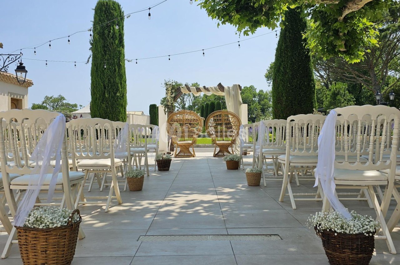 Allée centrale décorée pour une cérémonie en extérieur, entourée de chaises blanches et de paniers fleuris.