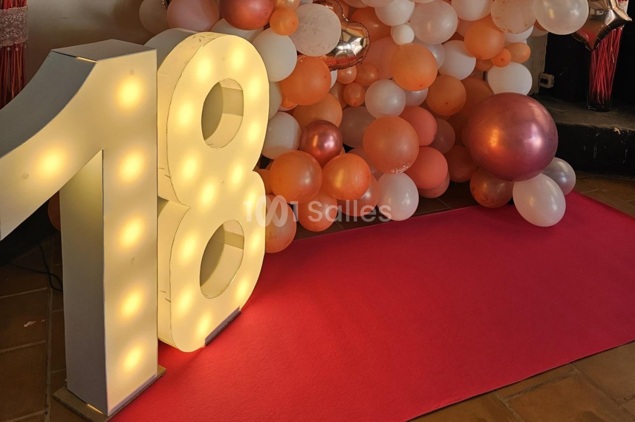 Chiffre 18 lumineux devant un mur de ballons roses, blancs et orange, avec un tapis rouge au sol.