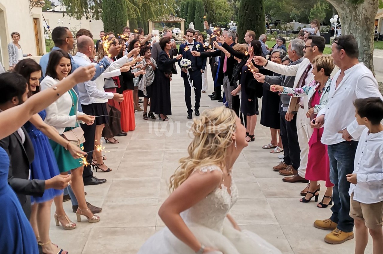Une mariée court vers un groupe de personnes tenant des cierges magiques, formant une allée festive en extérieur.