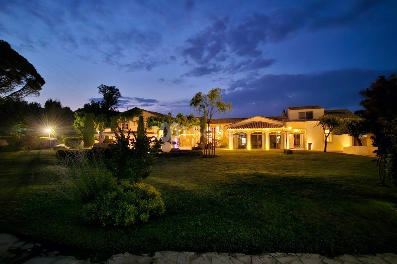Grande maison éclairée entourée de végétation et d'un jardin, photographiée au crépuscule sous un ciel nuageux.
