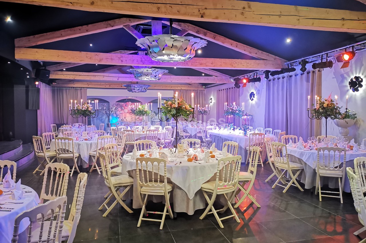 Salle de réception décorée avec des tables rondes, nappes blanches, chandeliers et arrangements floraux.
