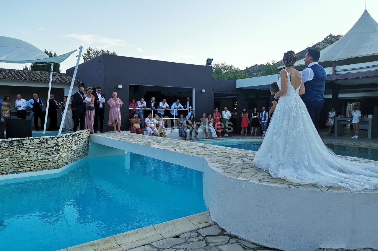 Un couple de mariés debout près d'une piscine, entouré d'invités observant la scène en extérieur.