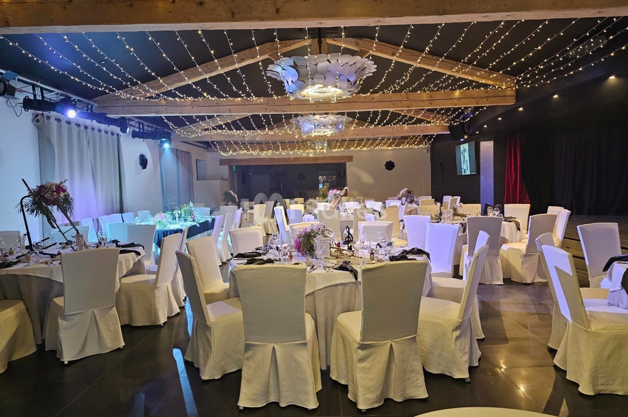 Salle de réception décorée avec des tables rondes dressées, des chaises blanches et un plafond illuminé de guirlandes…