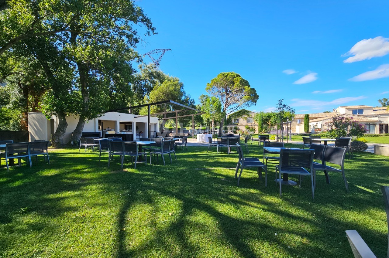 Espace extérieur avec tables et chaises sur une pelouse, entouré d'arbres et de bâtiments sous un ciel bleu.