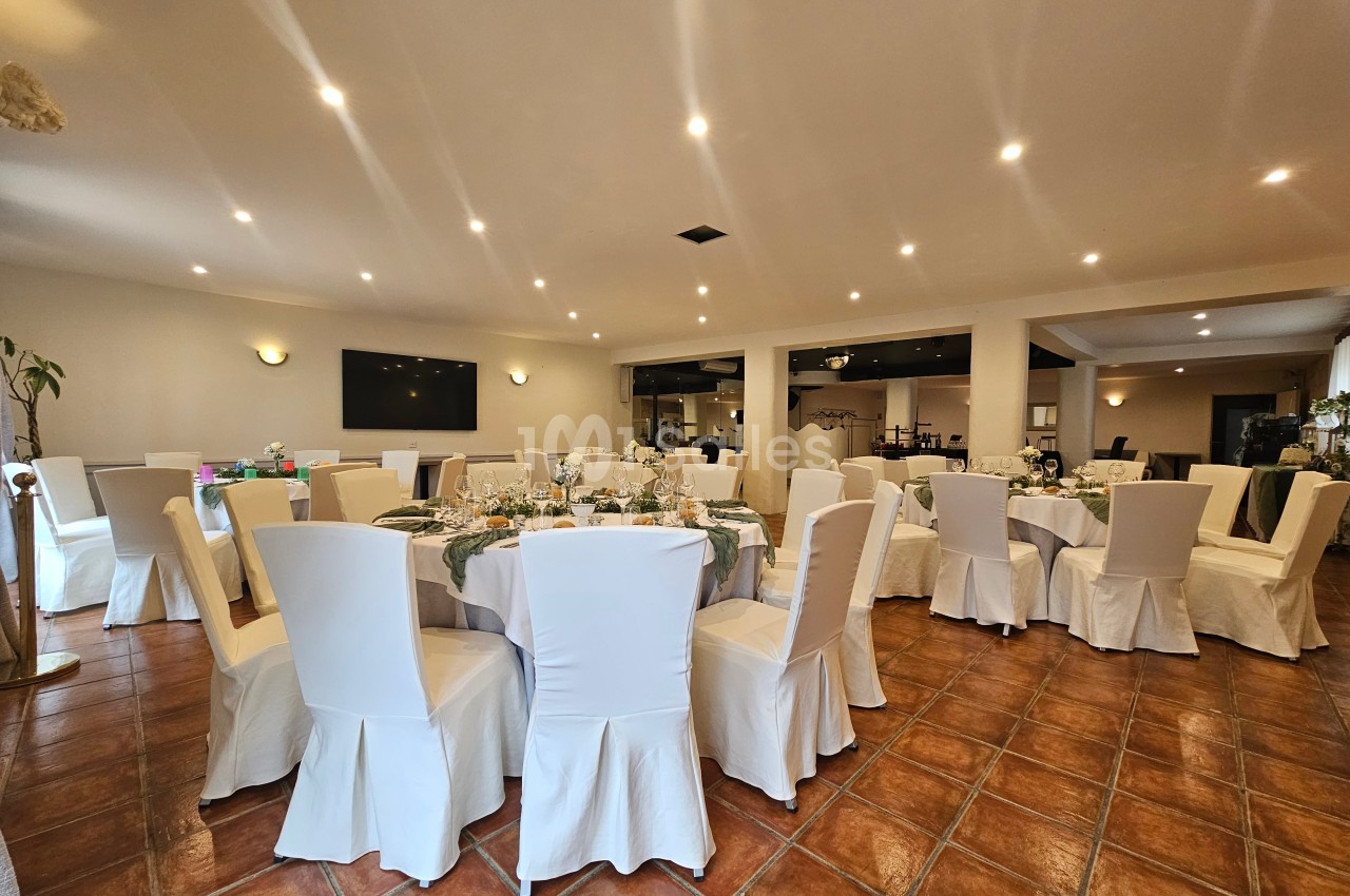 Salle de réception lumineuse avec des tables rondes dressées, entourées de chaises blanches et un sol en carrelage.