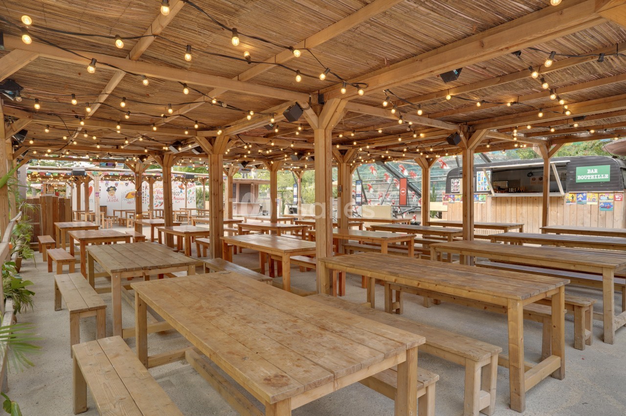 Terrasse couverte en bois avec tables et bancs, éclairée par des guirlandes lumineuses, dans un cadre extérieur.