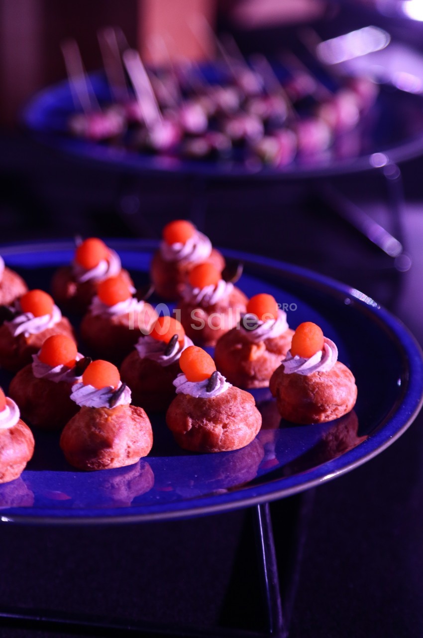 Plateau de petits choux garnis de crème et décorés de billes orange, avec des amuse-bouches flous en arrière-plan.
