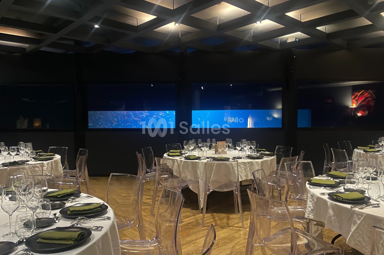 Salle de restaurant élégante avec tables dressées, nappes blanches et vue sur de grands aquariums éclairés.